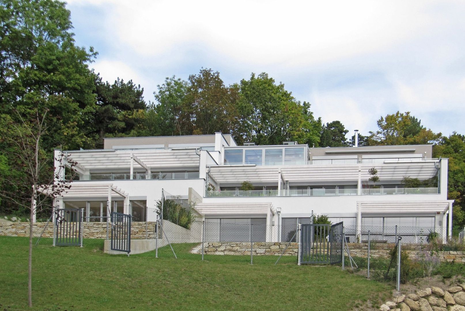 Foto einer modernen, terrassierten Wohnhausanlage in Hanglage mit weißen Fassaden, großen Fensterfronten und Glasbalkonen vor einer bewaldeten Kulisse.
