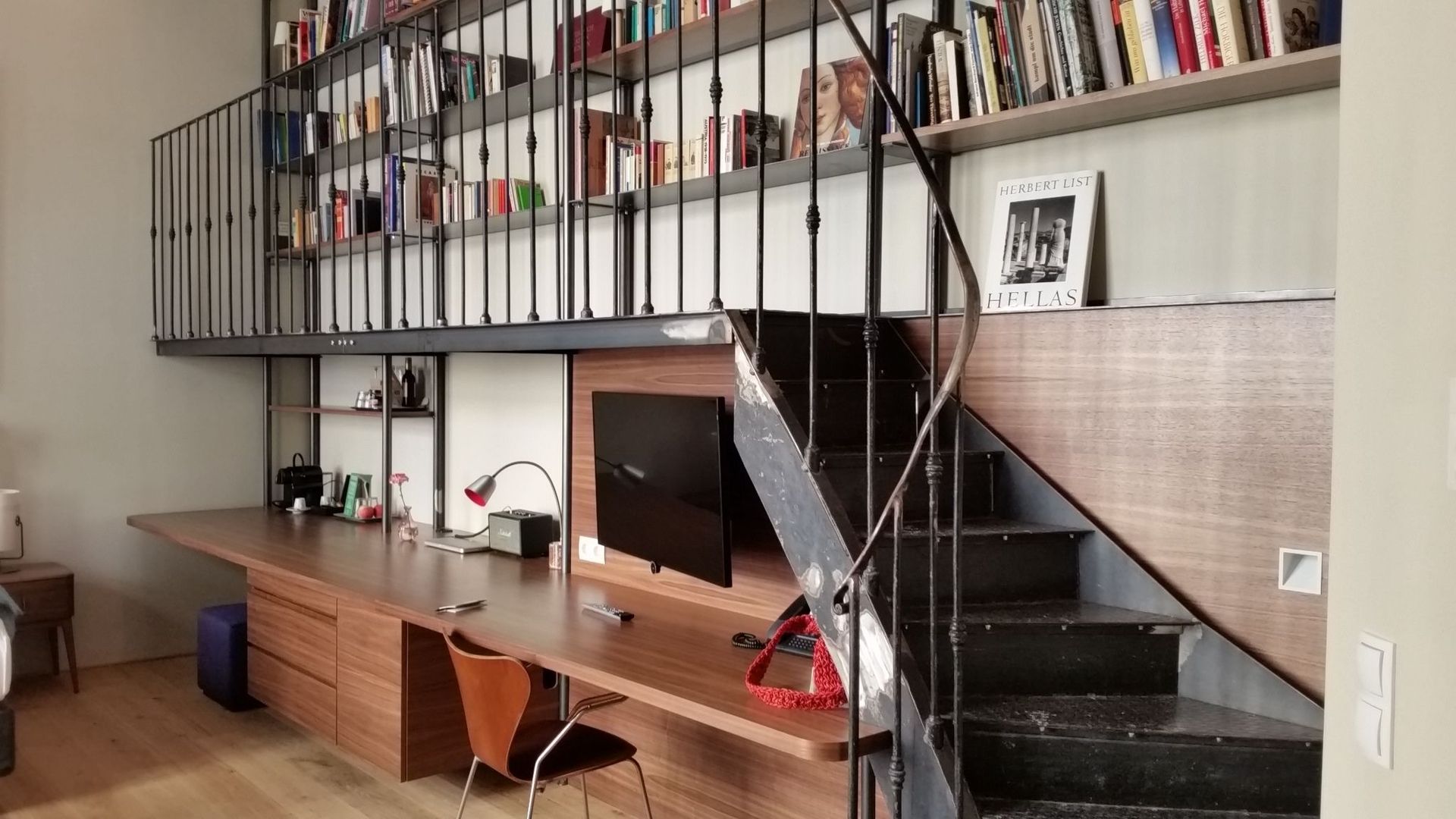 Moderner Arbeitsbereich mit langem Holzschreibtisch unter einer Galerie-Bibliothek, die über eine Metalltreppe erreichbar ist.