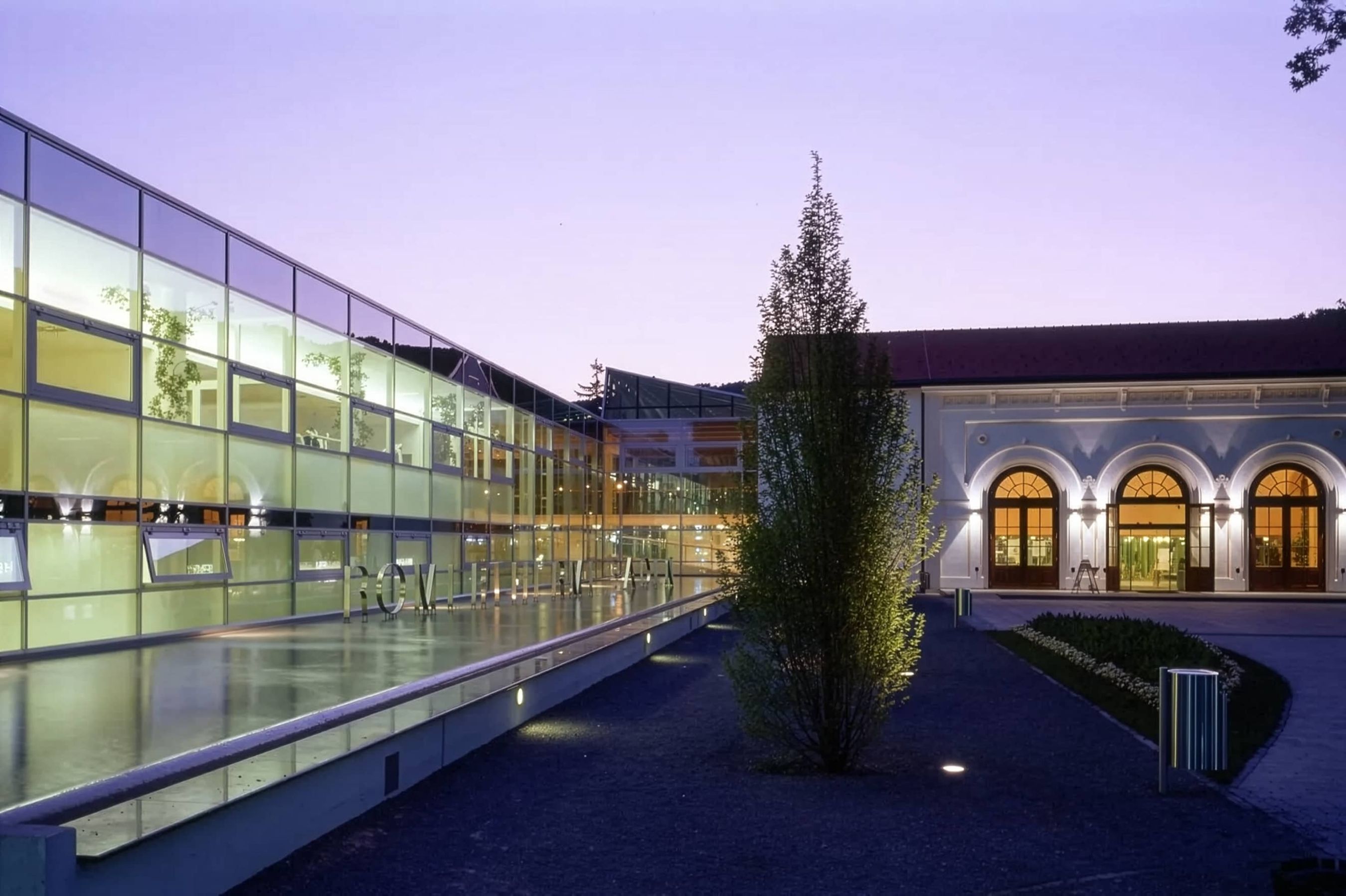 Abendaufnahme eines beleuchteten Architektur-Ensembles aus einem modernen Glaskomplex mit Wasserbecken und einem historischen Gebäude mit Rundbogenportalen.