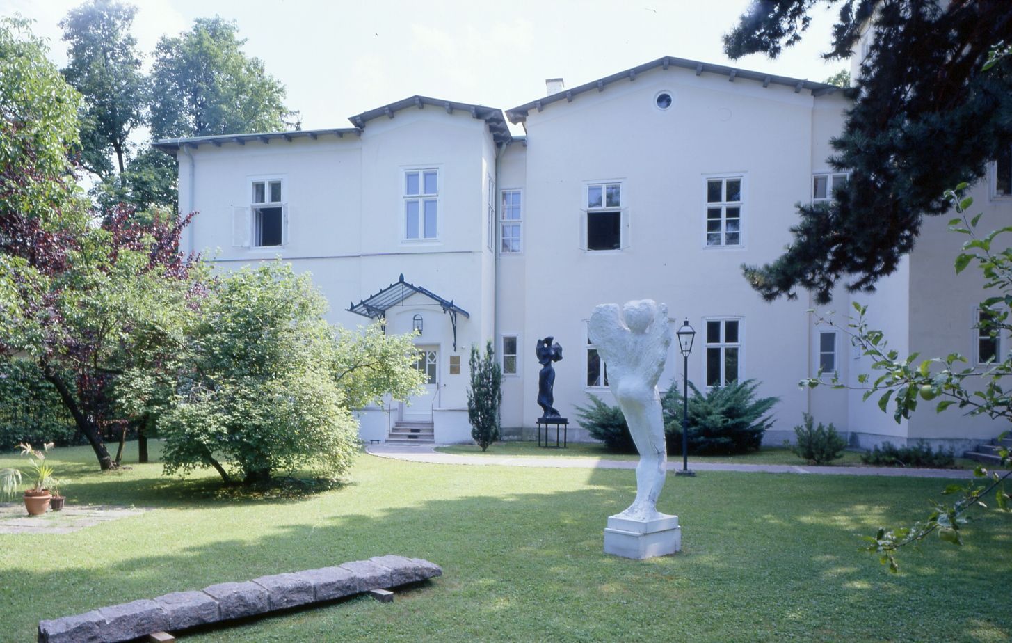 Außenansicht eines revitalisierten, hellen Gebäudes mit klassischer Fassade, gelegen in einer gepflegten Gartenanlage mit Rasenflächen und mehreren modernen Skulpturen.