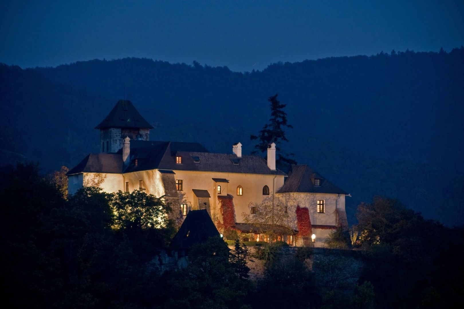 Nachtaufnahme der beleuchteten Burg Oberranna inmitten dunkler Bewaldung vor einer hügeligen Silhouette.