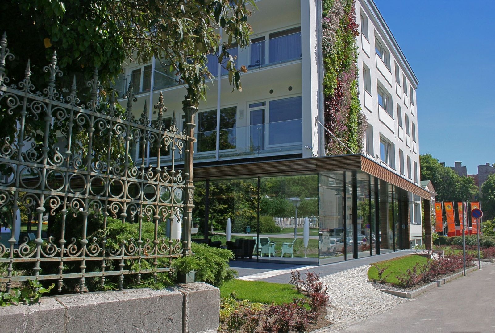 Modernisierte Fassade des Hotels At The Park mit verglastem Erdgeschoss, vertikaler Begrünung und historischem Schmiedeeisenzaun im Vordergrund.