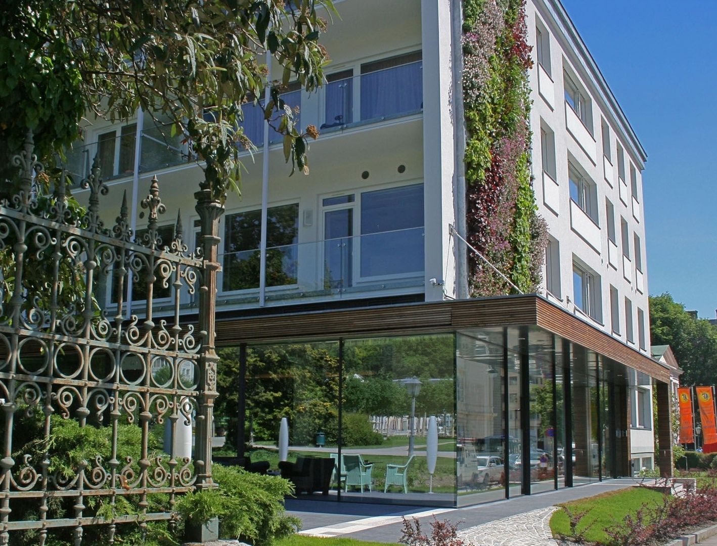 Modernisierte Fassade des Hotels At The Park mit verglastem Erdgeschoss, vertikaler Begrünung und historischem Schmiedeeisenzaun im Vordergrund.