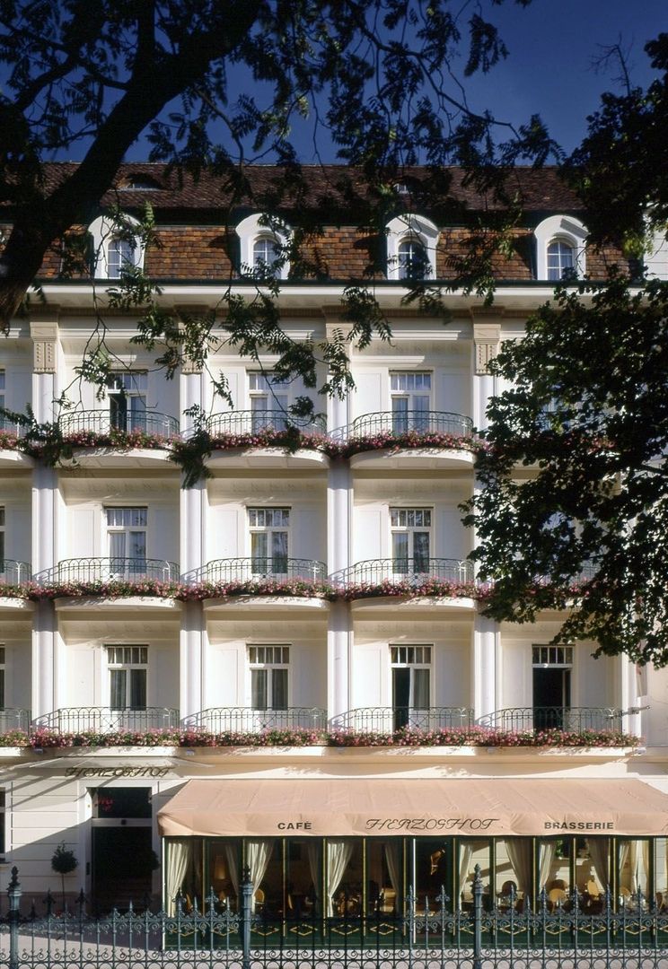 Fassade des Hotels Herzoghof in Baden mit blumengeschmückten Balkonen und dem Außenbereich von Café und Brasserie.