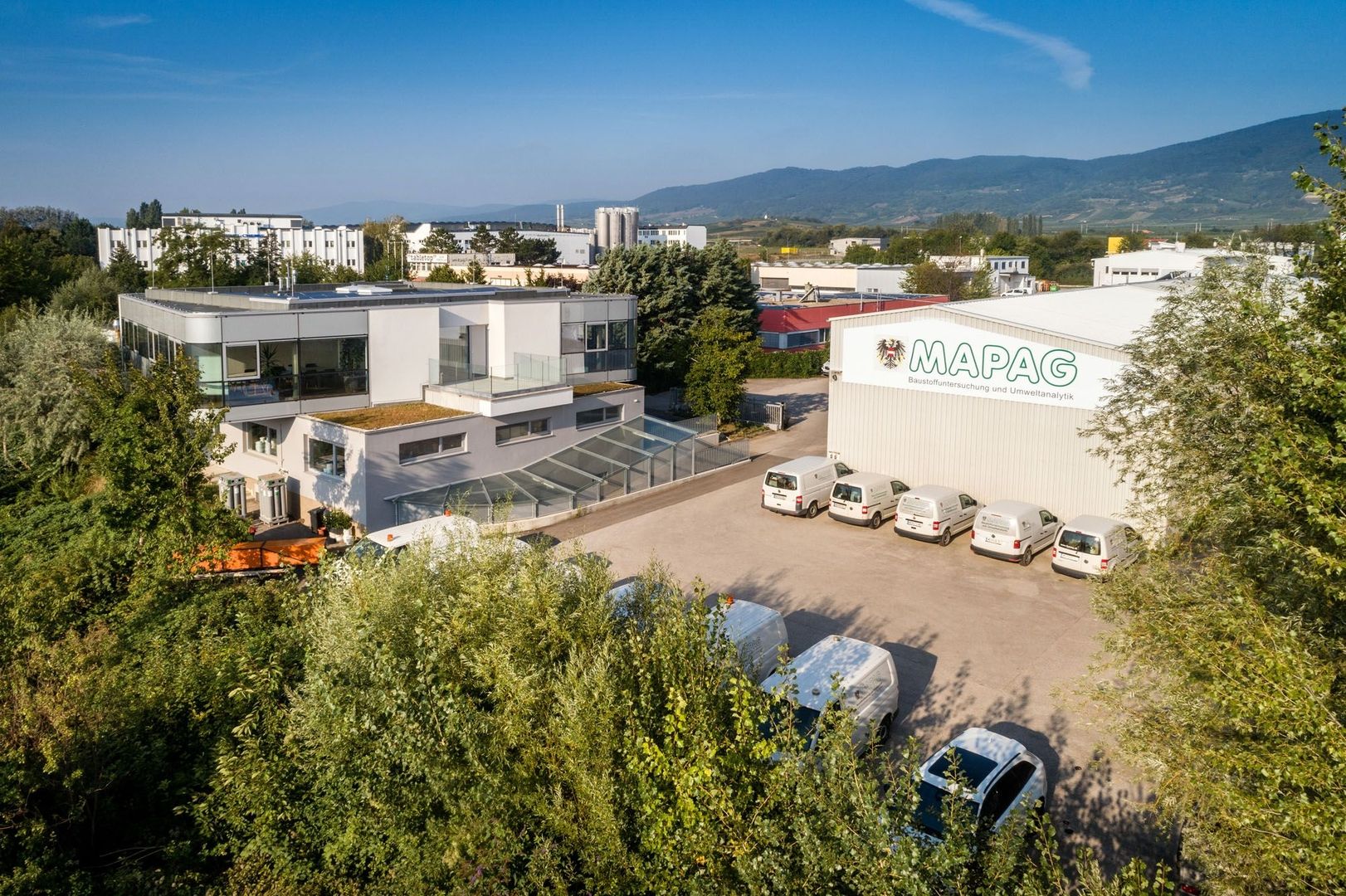 Luftaufnahme des Mapag-Firmengeländes mit modernem Labor-Anbau, Lagerhalle und Firmenfuhrpark vor einer Hügellandschaft.