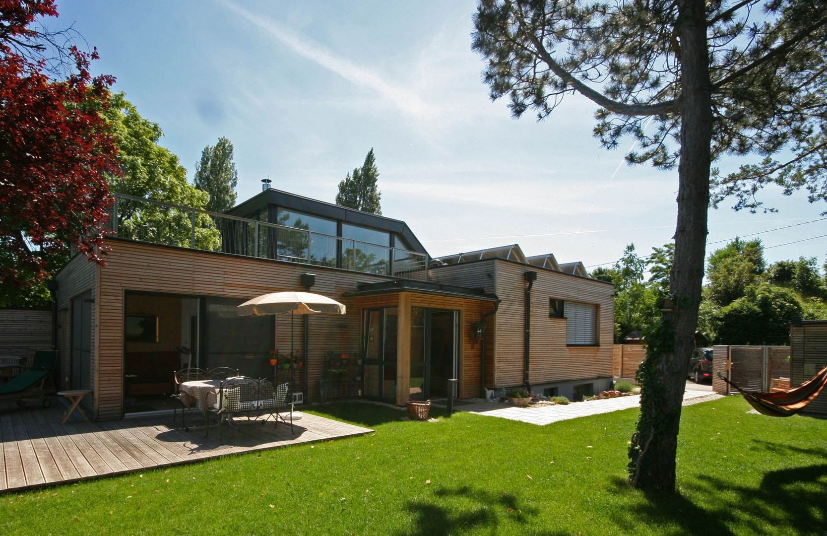 Moderner Holzbau mit Flachdach, großzügiger Dachterrasse und einer vorgelagerten Holzterrasse inmitten eines grünen Gartens.