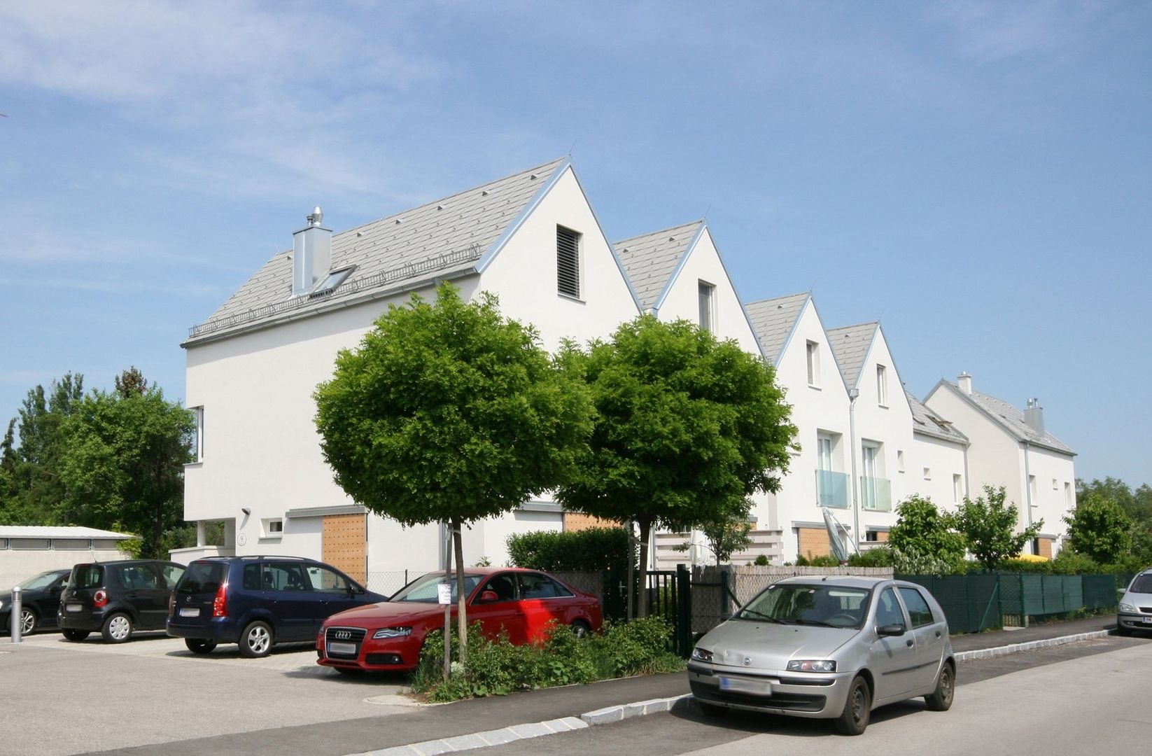 Foto einer modernen Reihenhausanlage mit weißen Fassaden und grauen Satteldächern, davor geparkte Autos und kugelförmig geschnittene Bäume an einer Straße.