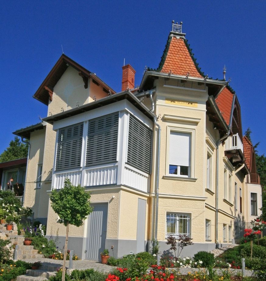 Historische Villa mit neuem, weißem Glasanbau und markantem ziegelgedeckten Ecktürmchen vor blauem Himmel.