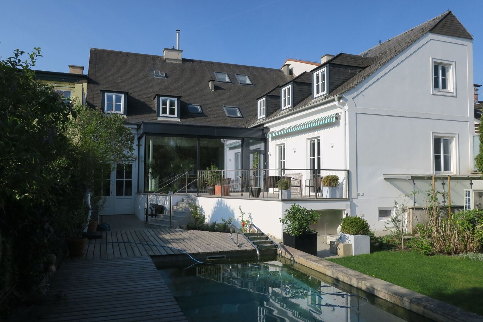 Modernisierte weiße Villa mit flach gedecktem Glas-Zubau, erhöhter Terrasse und einem Naturpool im Garten.